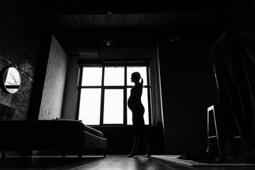Silhouette of a pregnant woman in a black dress near the window. Black and white photo of a pregnant woman by the window.