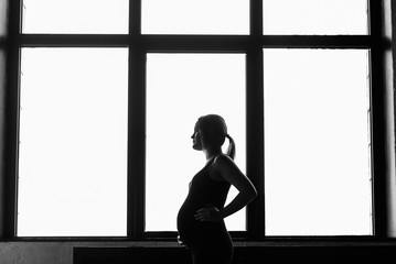 Silhouette of a pregnant woman in a black dress near the window. Black and white photo of a pregnant woman by the window.