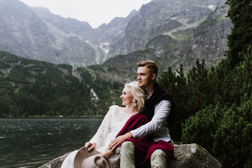 Beautiful bride in a boho style dress and groom cuddle m sit near the lake on a background of mountains. Wedding photo shoot in the mountains. Black and white photo of a bride and groom 