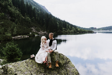 Beautiful bride in a boho style dress and groom hug and sit near the lake against the backdrop of the mountains. Wedding photo shoot in the mountains.