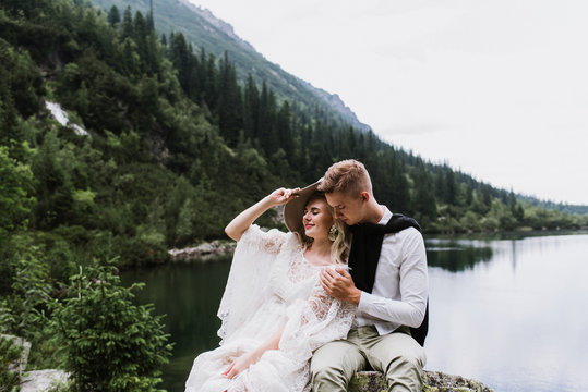 Beautiful bride in a boho style dress and groom hug and sit near the lake against the backdrop of the mountains. Wedding photo shoot in the mountains.