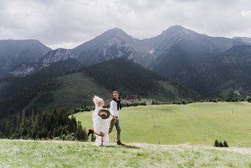 Naklejka premium Beautiful bride in a boho style dress and groom walk on the field near the mountains. Wedding photo shoot in the mountains.