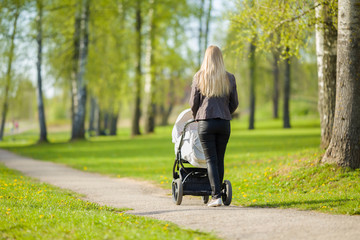 One young mother pushing white baby stroller and slowly walking at town green park in warm, sunny spring day. Spending time with infant and breathing fresh air. Enjoying stroll. Back view.