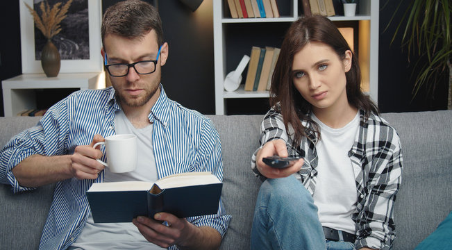 Spouses trying to divert themselves sitting on sofa, woman changing TV channels while man reading book