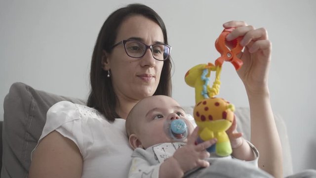  Caucasian young mother brunette and funny newborn baby boy playing with stuffed animal toys while sitting on the bed in the living room. Slow-motion. Portrait of mom and child.