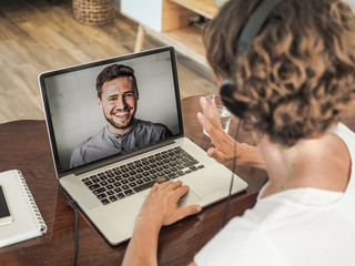 woman having a video conference call with a man waving into her laptop wearing a headset with...