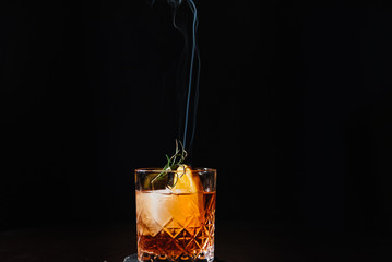 Whiskey with orange, ice and smoking rosemary close-up.