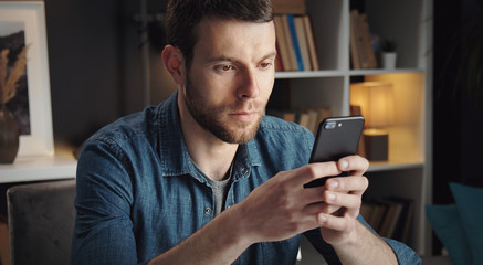 Obraz na płótnie Canvas Cropped shot of young man in denim shirt holding smartphone and texting staying in apartment