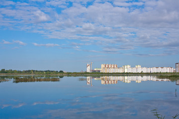 A clean lake with reeds, a blue cloudy sky and the construction of high-rise buildings in the background.