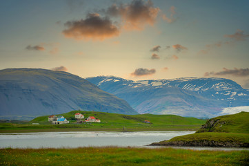 Fototapeta premium The house and ranch are located on a green meadow next to the lake. The background is high mountains, the sunset and beautiful sky, In the countryside in Iceland.