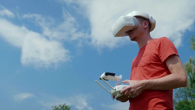 Man controls a quadrocopter through a remote control and looks at the video with goggles on his head. Drone operator in red T-shirt in a park shoots video on drone. Blue sky on the background
