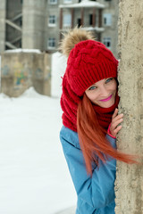 Portrait of young beautiful woman in red hat and red comforter hiding behind grunge concrete wall 