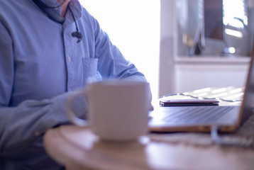 Man working from home in blue shirt at kitchen table with laptop computer and smart phone