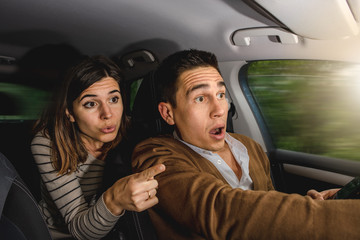 Caucasian couple inside car in action with blurred exterior background. Man driving while woman is pointing with finger something on the road. Astonishment, surprised and upset looking ahead.