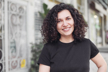 Happy woman with diastema, dark curly hair smiling and laughing. Looking at the camera, outside on...