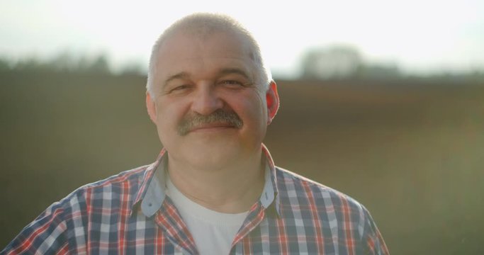 Slow motion: Close up of the senior Caucasian good looking wise man farmer in a hat looking at the side, turning face to the camera and smiling in the wheat field. Portrait