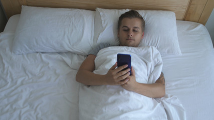 Young handsome man lies in bed uses the phone. Top view