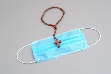 Prayer during a pandemic. Medical mask and wooden catholic cross with rosary on gray background.