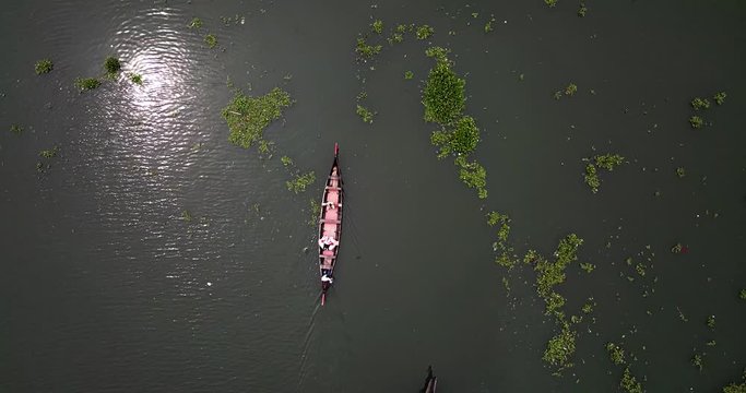 Aerial track shot with top down view of single traditional motorized wooden boat floating across natural lake with aquatic plants and reflecting sunlight on the surface, Kerala, India.