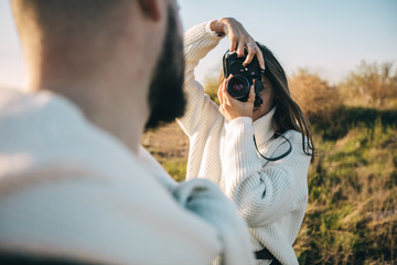 A guy and a girl love each other, smile, hug, kiss, laugh, enjoy life in the forest on a cliff, in the grass. Girl holding a film camera in her hands, photographs a guy, sunset in the background.
