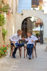 Two cute boys are talking sitting on wooden chairs. Boys imitate parents to businessmen. The boys are sitting on chairs with their legs crossed and beating with fists.