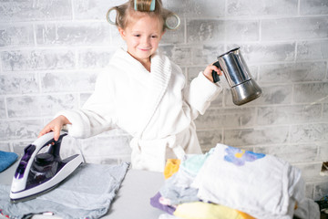 a little happy girl in a white coat with curlers on her head on a light background holds a coffee maker in her left hand, and in her right hand holds an iron on a blue towel. horizontal position