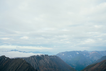 Atmospheric minimalist alpine landscape with big rockies and giant snowy mountains. Low cloud above beautiful rocky ridge. Wonderful tranquil highland scenery. Flying over mountains above clouds.