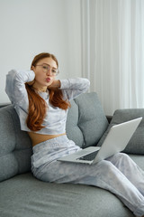 A red-haired girl in pajamas and glasses sits half-turn on the couch with a laptop on her lap and sends a kiss.