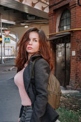 Naklejka na ściany i meble Portrait of a girl with long hair in a coat and a backpack standing on a street in St. Petersburg, a brick old building and bridge beams located close to a residential building
