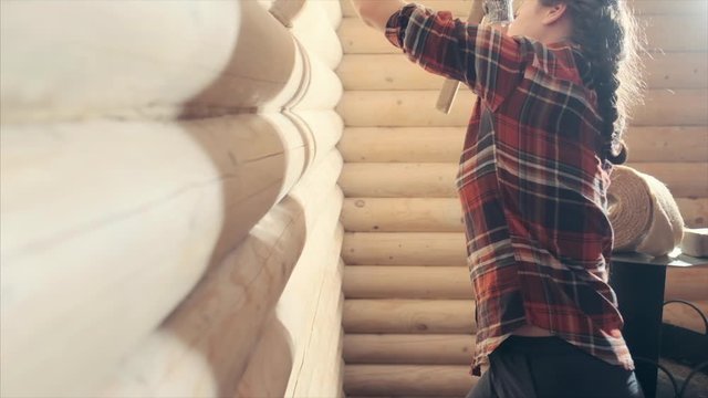 Young woman caulking and sealing wooden logs with oakum. Pretty woman works inside the wooden building and operates with hammer. Young feminist girl worker. Gender equality concept