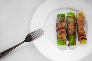 Three celery stalks, wrapped in bacon and toasted, lie on a white plate. To the left of the plate is a fork