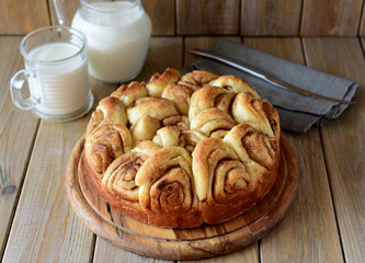 Delicious yeast cake with cinnamon on a wooden platter in the foreground, a jug of milk and a glass of milk in the background on a wooden table