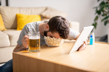 Obraz na płótnie Canvas Tired man sitting at coffee table and sleeping in popcorn bowl while watching video on tablet at home, isolation period concept