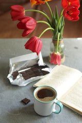 On the table is a cup of coffee, chocolate bar, a book and flowers.
