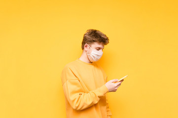 A young man in casual clothes and a protective gauze mask stands on a yellow background and uses the Internet on a smartphone. Coronavirus pandemic. COVID-19. Quarantine.