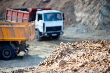 Pile of ore rocks over out of focus background with two heavy multi-ton heavy mining trucks exporting minerals from open-pit mine. Concept of shipping and protecting the environment and nature.