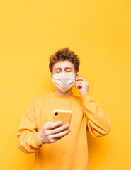 Positive guy in a medical mask with a smartphone in his hands listens to music in wireless headphones with closed eyes on a yellow background. Vertical. Coronavirus pandemic. COVID-19. Quarantine.