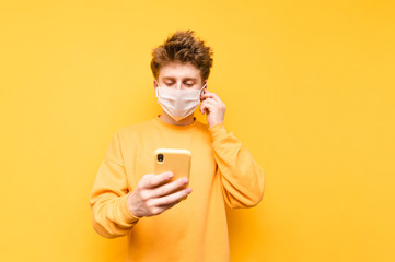Positive young man in a gauze protective mask and. wireless headset listens to music on a yellow background and uses a smartphone. Coronavirus pandemic. COVID-19. Quarantine.