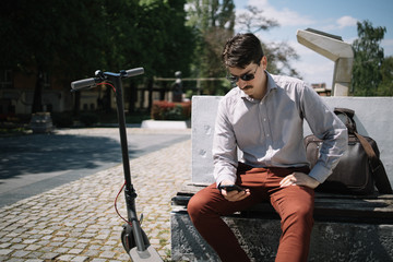 Man with scooter sitting in city center. Happy man looking at his smartphone while sitting on a bench next to scooter in city centre. © Synergic Works OÜ