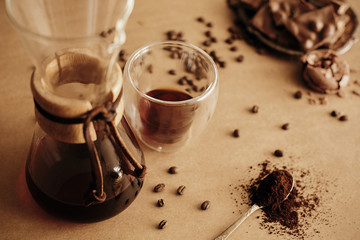 Glass cup with fresh coffee, roasted beans, grounded coffee on spoon, glass flask for filter coffee and macarons, chocolate and cinnamon on brown background. Coffee Essentials.