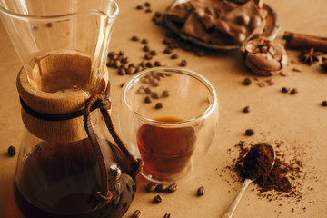 Making filter coffee in glass flask. Glass cup with fresh coffee, roasted beans, grounded coffee on spoon and macarons, chocolate and cinnamon on brown background. Coffee Essentials