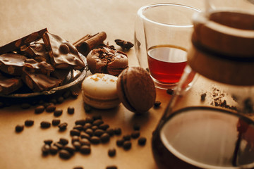 Coffee roasted beans, glass cup with fresh coffee, and macarons,chocolate, cinnamon on brown background with glass flask for filter coffee. Stylish image