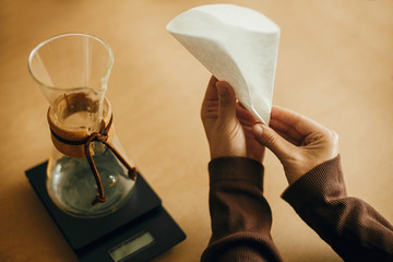 Hands folding paper filter for pour over and glass kettle on scale on brown background. Preparing for alternative coffee brewing v60. Fold coffee filter