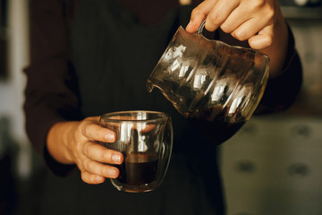 Professional female barista in black uniform making drip coffee. Person pouring fresh aromatic coffee from glass kettle in cup.  Alternative coffee brewing, v60.