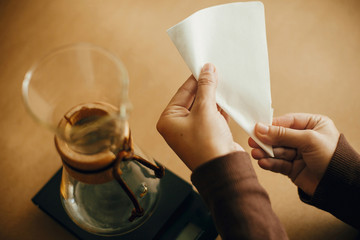 Preparing for alternative coffee brewing v60. Fold coffee filter. Hands folding paper filter for pour over and glass kettle on scale on brown background.