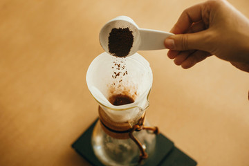 Hands pouring grounded coffee in filter. Preparing for alternative coffee brewing v60. Person holding spoon with grind coffee on background of glass kettle with pour over on scale