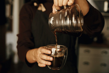 Professional female barista in black uniform making drip coffee. Person pouring fresh aromatic coffee from glass kettle in cup.  Alternative coffee brewing, v60.