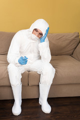 Sad man in hazmat suit and medical mask holding thermometer on sofa at home
