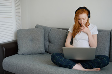 The red-haired girl sits on the sofa, putting a laptop on her legs and staring thoughtfully at it.