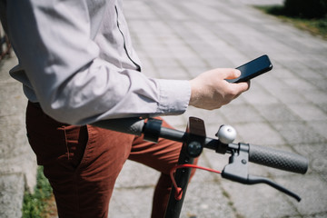 Cropped guy using phone while standing outdoor. Close-up of man's hand holding black mobile phone and leaning over scooter rudder.